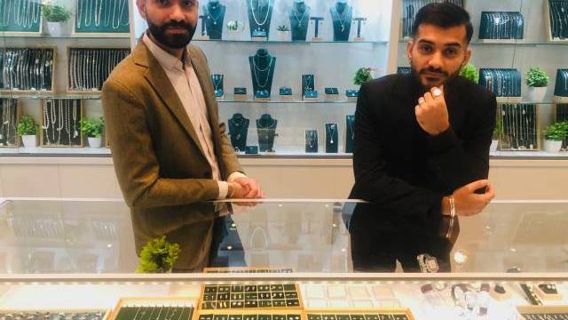 Två män står bakom en smyckesdisk i glas med ringar och accessoarer, med hyllor med halsband och smycken i bakgrunden.