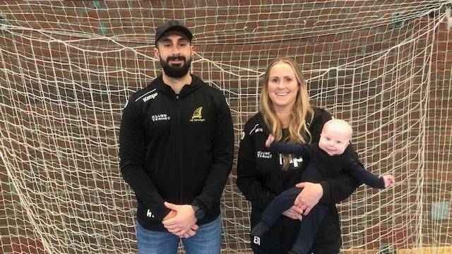 Två vuxna i sportkläder står framför ett inomhusfotbollsmål; kvinnan till höger håller i en bebis.