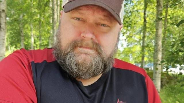 En skäggig man med brunvit keps och svartröd skjorta sitter utomhus i ett skogsparti där solljuset silar genom träden.