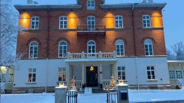 En trevåningsbyggnad i tegel och stuckatur är upplyst med utomhusbelysning, marken är täckt av snö och vid entrén står små dekorerade träd.