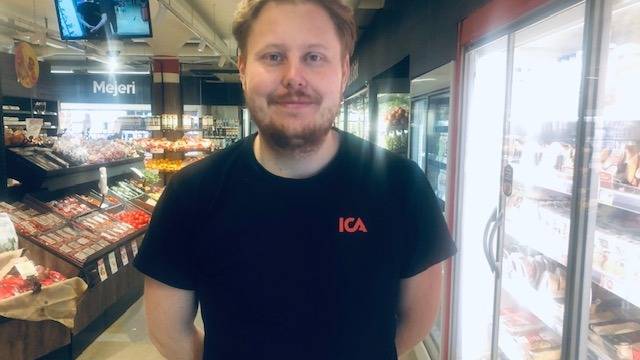 En man med en ICA-t-shirt står i en mataffär i närheten av kyl- och frysdiskar och tittar in i kameran.