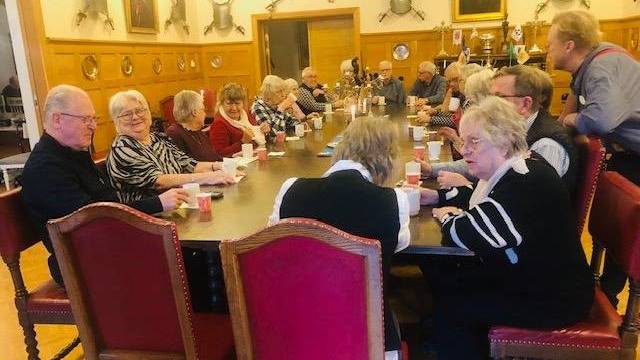 En grupp äldre människor sitter runt ett långt träbord i ett inrett rum, dricker ur koppar och samtalar.