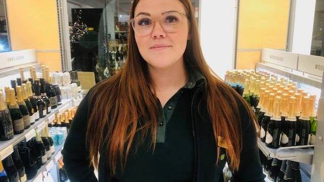 En kvinna med långt brunt hår och glasögon står i en butiksgång med flaskor med mousserande vin och champagne.