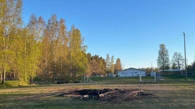 Ett gräsbevuxet fält med en hög av grenar i mitten, omgivet av träd och en vit byggnad i bakgrunden under en klarblå himmel.