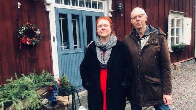 Två personer står utanför ett rött trähus med blå dubbeldörr, julkrans och vintergrönska vid entrén.