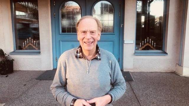 En äldre man i grå tröja står leende framför blå dubbeldörrar med glaspartier och två fönster på vardera sidan.