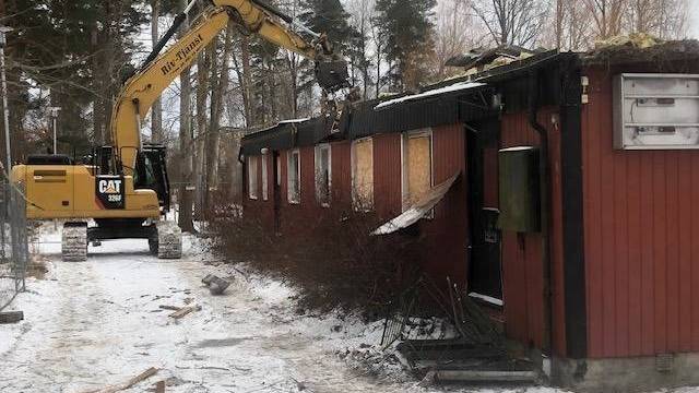 En gul grävmaskin river en röd träbyggnad på en snöig mark, med bråte utspridd i närheten och träd i bakgrunden.