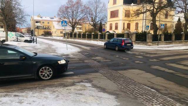 Bilar kör på en delvis snötäckt gata i ett bostadsområde med stora hus och kala träd en molnig dag.