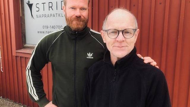 Två män står utomhus framför en röd byggnad med en klinikskylt i bakgrunden. Den ena mannen har skägg och bär en grön jacka, den andra har glasögon och en svart jacka.