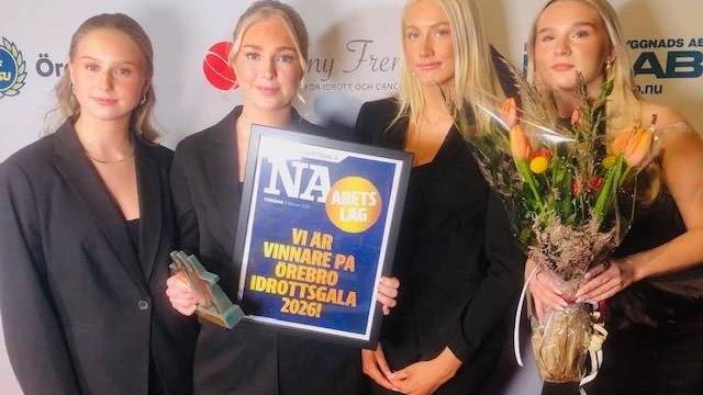 Fyra formellt klädda kvinnor står tillsammans och håller i en pokal, en blombukett och en skylt med texten 