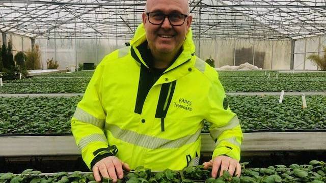 En man i en ljusgul jacka står i ett växthus och ler med händerna på rader av gröna bladväxter.
