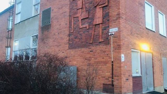 En röd tegelbyggnad med en reliefskulptur av två abstrakta människofigurer på väggen ovanför en dörröppning, bredvid några buskar och en tänd ytterbelysning.