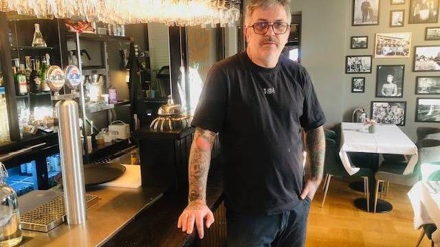 En man med glasögon och tatueringar står bakom disken i en restaurang eller bar, med bord, stolar och inramade foton på väggen i bakgrunden.