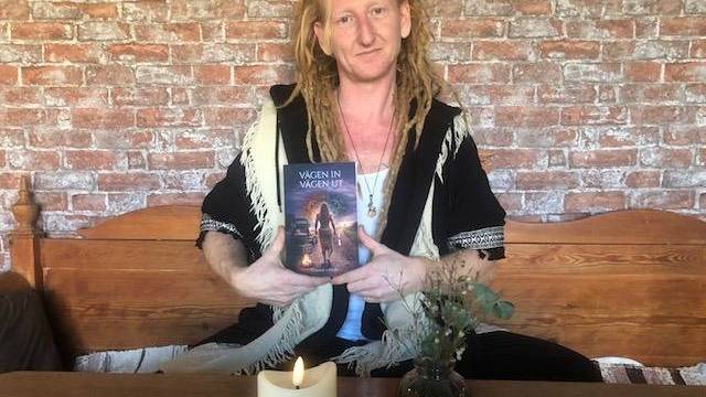 En person med långa, blonda dreadlocks sitter på en träbänk och håller i en bok. Ett tänt ljus och en liten vas står på bordet framför dem; tegelvägg och inramade bilder i bakgrunden.