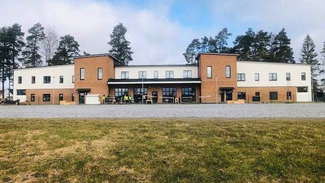 En modern tvåvåningsbyggnad med vit och brun tegelfasad, stora fönster och omgiven av träd, sedd från en gräsyta.