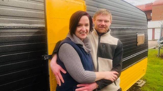 Ett par står och ler och omfamnar varandra framför en svartgul husbil på en gräsmatta.