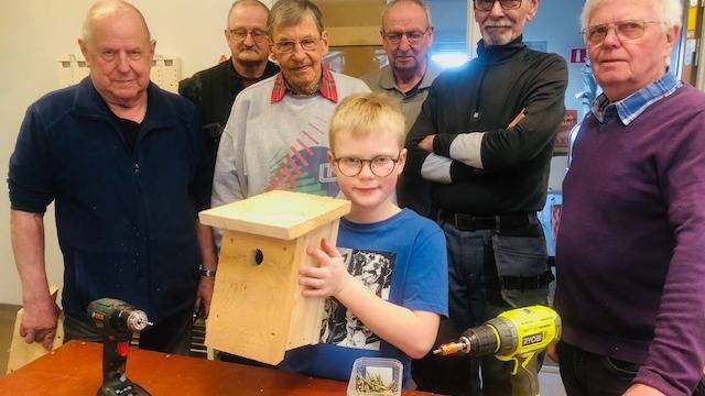 En ung pojke håller i en fågelholk av trä, omgiven av sex äldre män i en verkstad. På bordet står två borrmaskiner och en låda med skruvar.