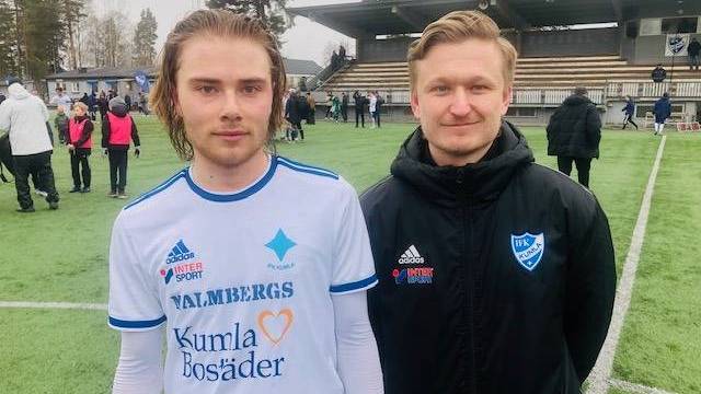 Två män står på en fotbollsplan; den ene bär en vit fotbollströja, den andre en svart jacka. Det finns människor och läktare i bakgrunden.
