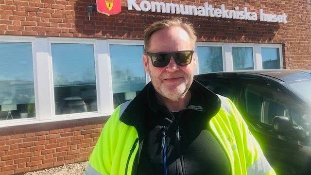 En man iförd solglasögon och varseljacka står framför en tegelbyggnad med texten 