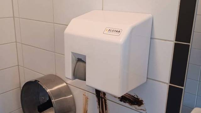 En vägg på en offentlig toalett med handtork och toalettpappersbehållare är nedsmutsad med en brun substans. Toalettpappersrullar ligger på en yta i närheten.