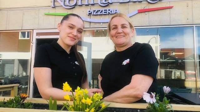 Två kvinnor står och ler utanför Nonna Bina Pizzeria, bakom en plantering med gula och vita blommor. Restaurangens skylt syns ovanför dem.