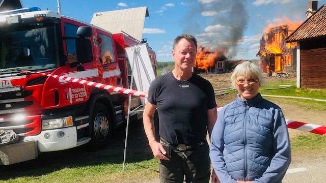 Två personer står framför en brandbil och ett avspärrat område, med ett hus som brinner i bakgrunden och rök som stiger upp mot himlen.