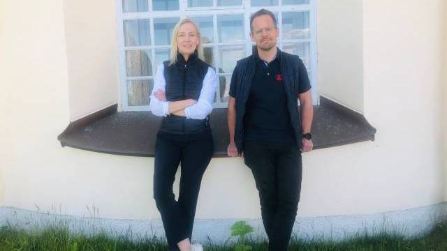 Två personer står utomhus framför ett stort fönster, lutade mot väggen med korsade armar och ben, på en gräsmatta.