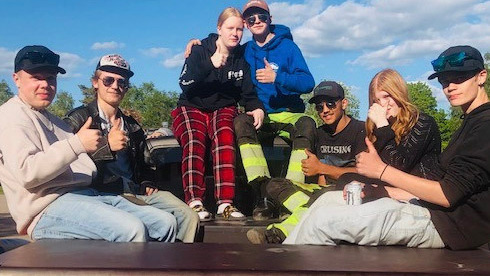 Sex ungdomar sitter utomhus, några gör tummen upp-gester, med träd och en blå himmel i bakgrunden.