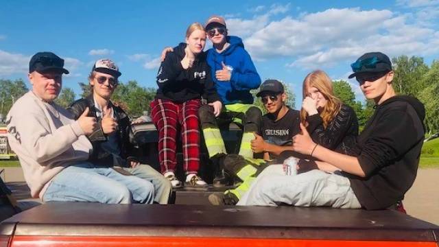 Sju personer sitter på baksidan av ett rött fordon, gör tummen upp och poserar för kameran en solig dag. Träd och en blå himmel syns i bakgrunden.