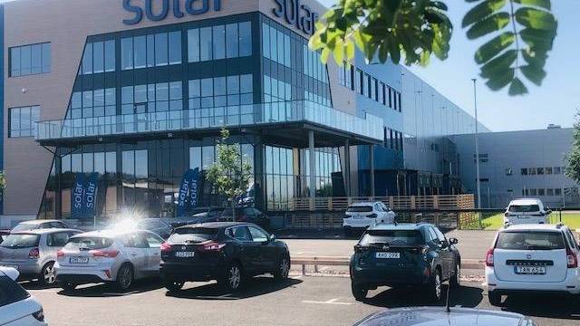 En modern kontorsbyggnad med skyltar med solmotiv, stora glasfönster och en parkeringsplats full av bilar en solig dag.