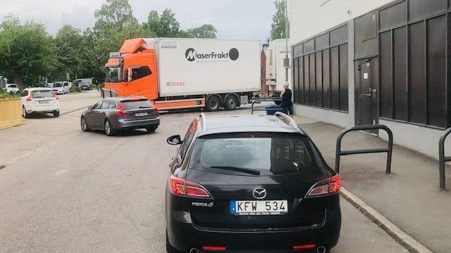 En svart Mazda med svenska registreringsskyltar står parkerad på gatan nära trottoaren, medan en lastbil och en grå sedan står längre fram, och en person står i närheten.