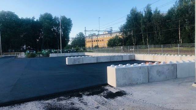En nyasfalterad yta med betongbarriärer, omgiven av träd och staket, med en gul byggnad i bakgrunden under en klar himmel.