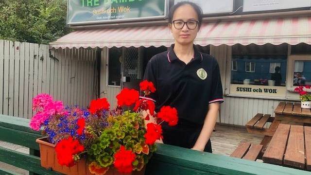En kvinna i svart uniform står vid ett grönt staket med en blomlåda framför en restaurang som heter Tre Bambu, och bakom henne syns uteserveringen.