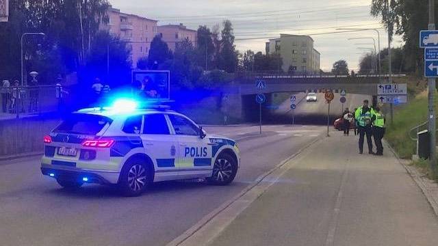 En polisbil med blå blinkljus spärrar av en väg nära en tunnel, medan två poliser står på trottoaren intill.