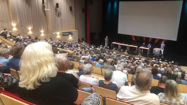 En stor publik sitter i en sal med teateruppställning vänd mot en scen där fyra personer sitter vid bord och en person står bredvid en skärm.