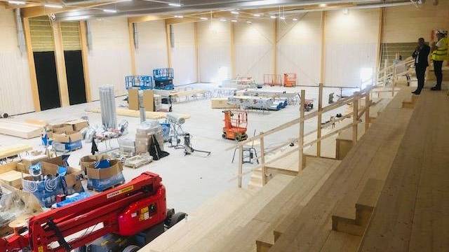 En stor byggarbetsplats inomhus med läktare av trä, utrustning, byggmaterial och två personer i skyddsutrustning som observerar från en högre nivå.