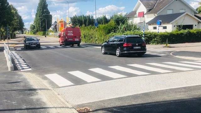 Tre fordon, däribland en röd skåpbil och två personbilar, kör på en asfalterad väg med övergångsställen i ett förortsområde en solig dag.