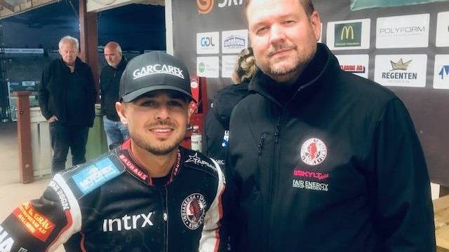 Två män står bredvid varandra inomhus; den ene har på sig en racerdräkt och en keps, den andre har på sig en svart jacka. Sponsorslogotyper syns på deras kläder och på väggen bakom dem.