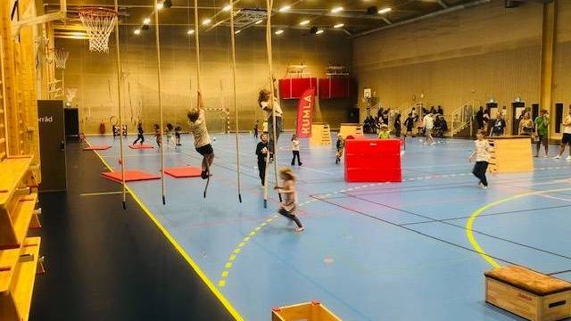 Barn leker och rör på sig på klätterrep, mattor och annan utrustning i en stor inomhushall, med människor i bakgrunden.