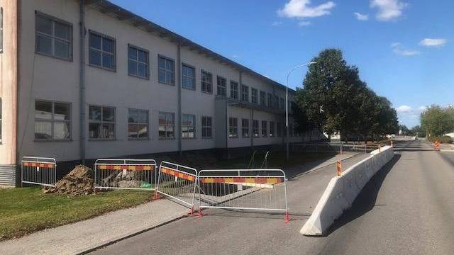 En byggstängsel spärrar av en del av trottoaren nära en byggnad, med trafikavspärrningar längs den intilliggande vägen under en blå himmel.
