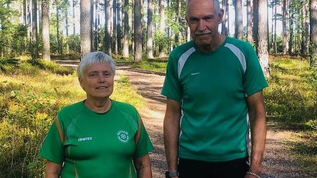 Två äldre personer står på en solbelyst skogsstig, båda klädda i gröna träningströjor. De är omgivna av träd och grönska.