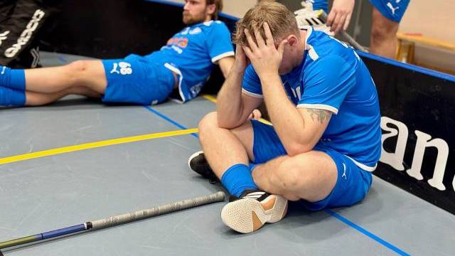 Två manliga innebandyspelare i blåa dräkter sitter på golvet, den ene med huvudet i händerna, och verkar upprörda efter en match. Innebandyklubbor syns bredvid dem.
