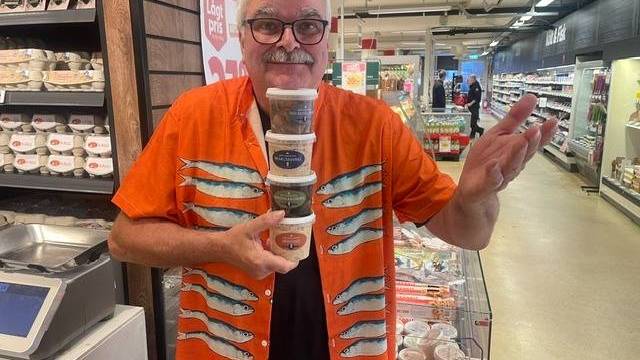 En man i en orange skjorta med fiskmönster står i en gång i en livsmedelsbutik med en stapel bestående av fem små matförpackningar i händerna och ler mot kameran.