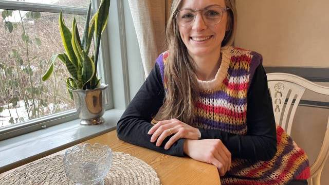 En kvinna med glasögon och en färgglad stickad väst sitter vid ett träbord vid ett fönster, med en glasskål och en krukväxt i närheten.