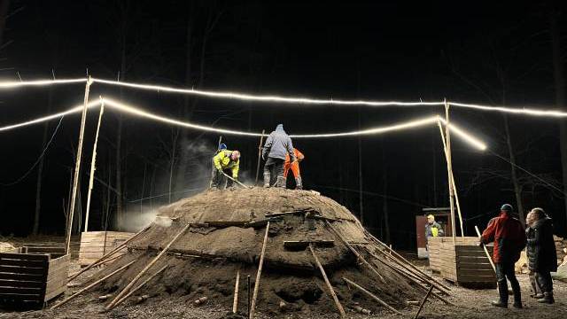 Flera personer arbetar på och runt en stor, kupolformad jordhög under starka lampor på natten, med träplankor och stolpar som stöd för strukturen.