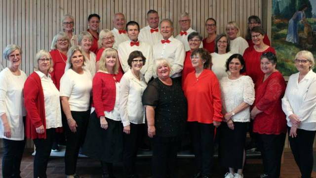 En grupp på 26 vuxna, de flesta kvinnor, står inomhus framför en trävägg. Några är klädda i rött, andra i vitt eller svart. Fyra män i mitten bär vita skjortor och röda flugor.