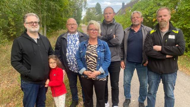 Sju vuxna och ett barn står tillsammans på en trädkantad stig och tittar mot kameran med uttryckslösa ansikten.
