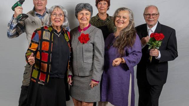 Sex vuxna står och poserar för ett gruppfoto mot en vanlig bakgrund; en håller i en borr, en annan håller i en bukett röda rosor och flera har färgglada kläder och leenden.