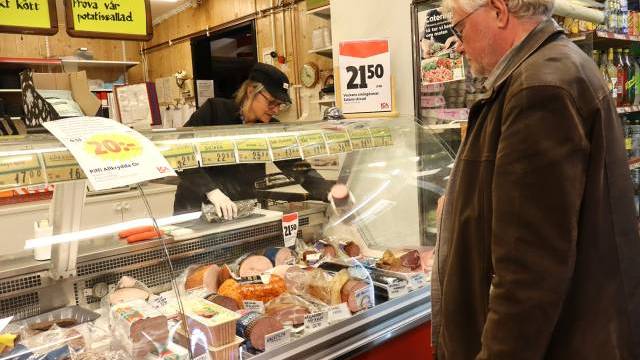 En man står framför en delikatessdisk med olika köttbitar medan en anställd bakom disken förbereder hans beställning i en livsmedelsbutik.