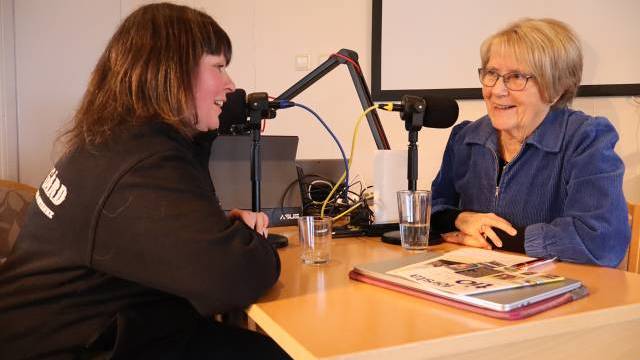 Två kvinnor sitter mitt emot varandra vid ett bord med mikrofoner, inspelningsutrustning, vattenglas och en tidning, vilket tyder på en podcast eller en intervju.